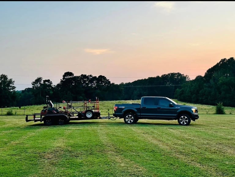 Blue pickup truck towing a farm trailer in an open green field at sunset with trees