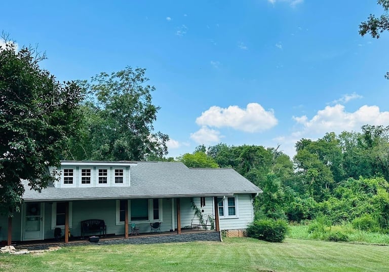 White farmhouse with green shutters and covered porch, surrounded by mature trees